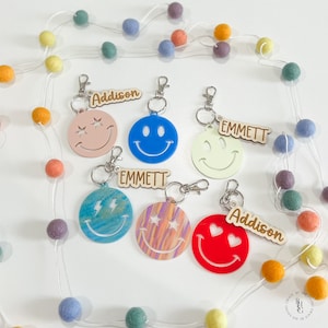 May include: Six colorful smiley face keychains with wooden name tags. The keychains feature different designs, including stars, lightning bolts, and hearts. The name tags read "Addison" and "Emmett".