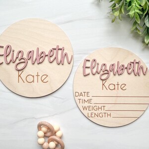 May include: Two circular light-colored wooden plaques featuring the name Elizabeth Kate in raised dusty pink script lettering. One plaque is plain, while the other includes engraved lines for date, time, weight, and length information.