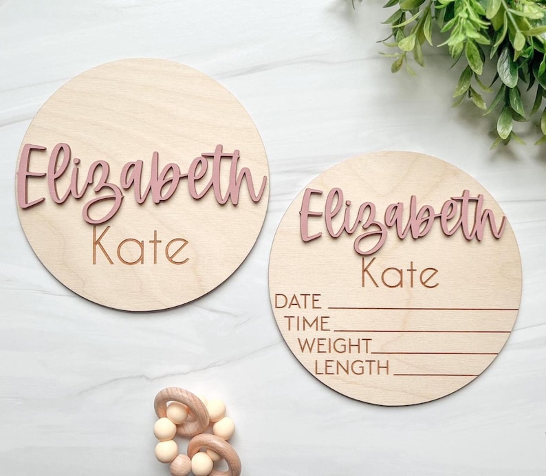 May include: Two circular light-colored wooden plaques featuring the name Elizabeth Kate in raised dusty pink script lettering. One plaque is plain, while the other includes engraved lines for date, time, weight, and length information.