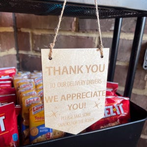 May include: A wooden sign with the words "THANK YOU TO OUR DELIVERY DRIVERS WE APPRECIATE YOU!" is hanging above a snack basket. The sign also says "PLEASE TAKE SOME SNACKS FOR THE ROAD!" The basket contains various snack packages.