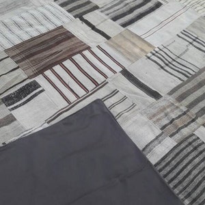 May include: A patchwork rug with a variety of neutral tones, including gray, beige, and brown. The rug is made of rectangular pieces with striped and solid patterns. A dark gray fabric is partially covering the rug.