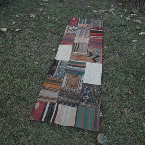 Puede incluir: Una alfombra larga y rectangular de patchwork con una variedad de colores y patrones. La alfombra está hecha de diferentes piezas de tela cosidas juntas, creando un diseño único y ecléctico. La alfombra está colocada sobre una superficie cubierta de hierba.