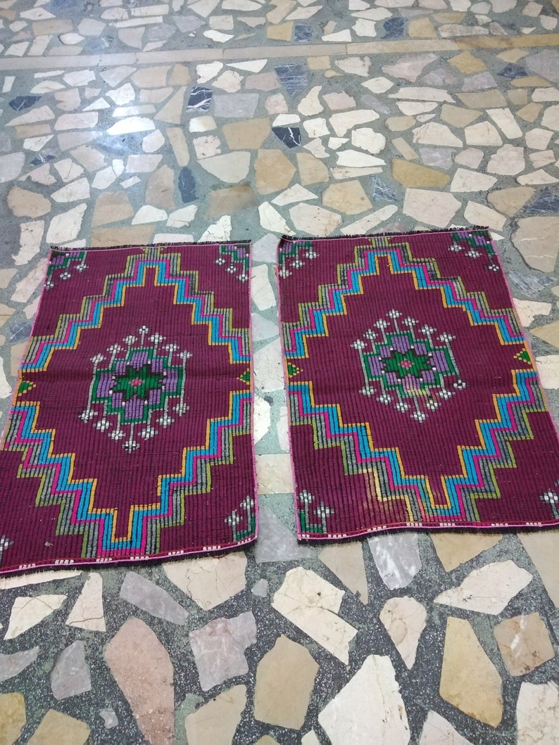 Puede incluir: Dos alfombras de color burdeos con intrincados patrones geom&eacute;tricos en tonos azules, verdes y rosas. Las alfombras tienen un borde con flecos y est&aacute;n colocadas sobre un suelo de piedra.