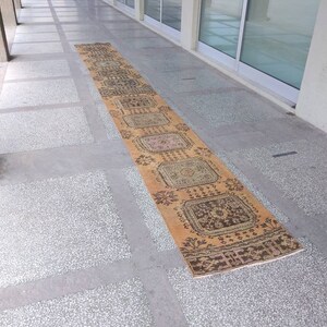 May include: A long, rectangular, beige and brown patterned rug with intricate designs. The rug is laid out on a tiled floor.