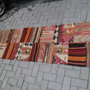 Op de afbeelding: Een lang, rechthoekig tapijt gemaakt van patchwork stof. Het tapijt is gemaakt van verschillende gekleurde stroken stof, waaronder rood, oranje, bruin en wit. Het tapijt heeft een traditionele, handgeweven uitstraling.