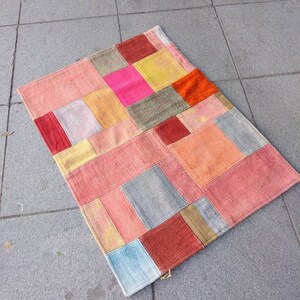 May include: A patchwork rug with a geometric design in shades of pink, red, orange, yellow, blue, and grey. The rug is made of woven fabric and has a fringed edge.