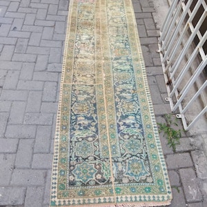 May include: A long, rectangular rug with a faded, vintage look. The rug has a cream background with a blue and green floral pattern. The rug has a fringe on both ends.
