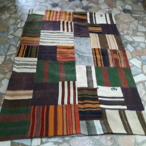 Może przedstawiać: Prostokątny dywan patchworkowy z różnorodnością kolorów i wzorów. Dywan składa się z licznych prostokątnych łatek w odcieniach brązu, zieleni, pomarańczy, bieli i fioletu. Niektóre łatki mają paski, inne są jednolite.