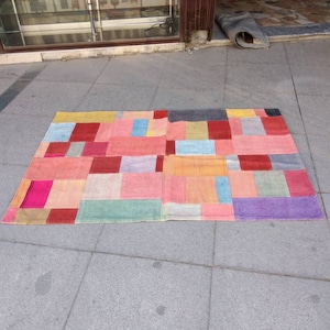 Könnte beinhalten: Ein bunter Patchwork-Teppich mit Quadraten aus rotem, rosa, blauem, gelbem, grünem und violettem Stoff. Der Teppich liegt auf einem grauen Fliesenboden.