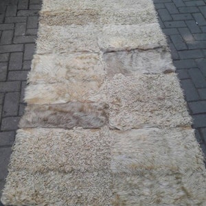 Könnte beinhalten: Ein rechteckiger, cremefarbener Schaffellteppich, der aus mehreren rechteckigen Teilen zusammengenäht ist. Der Teppich hat ein zotteliges, strukturiertes Aussehen mit verschiedenen Creme- und hellbraunen Farbtönen. Er liegt auf einer grauen Ziegeloberfläche.