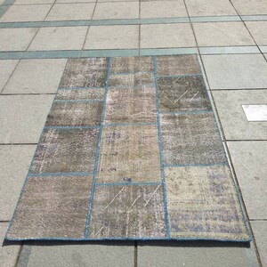 Puede incluir: Una alfombra de patchwork con un borde azul y una variedad de cuadrados beige y marrón. La alfombra está hecha de tejido tejido y tiene un aspecto desgastado.