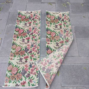 Op de afbeelding: Twee tapijten met een bloemenpatroon op een crème achtergrond en roze, groene en bruine bloemen. De tapijten liggen op een grijze tegelvloer.