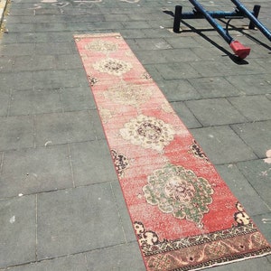 Puede incluir: Una alfombra larga y rectangular con un patrón rojo, rosa y crema. La alfombra tiene un diseño floral con detalles intrincados. La alfombra está colocada sobre una superficie de hormigón gris.