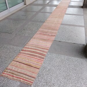May include: A long, narrow, handwoven rug with horizontal stripes in shades of pink, beige, brown, and yellow. The rug is placed on a gray stone surface, and appears to be made of natural fibers. The rug is a runner style.