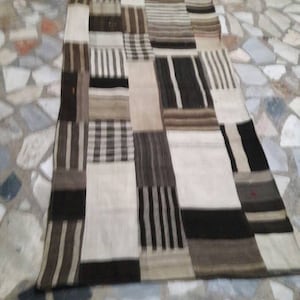 Może przedstawiać: Dywan patchwork z geometrycznym wzorem czarnych, białych i brązowych pasków. Dywan jest wykonany z tkanego materiału i ma rustykalny, ręcznie robiony wygląd.