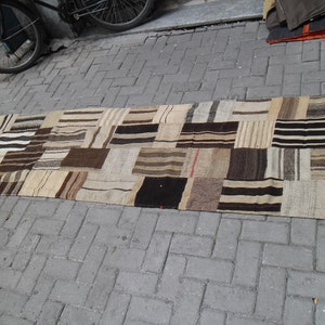 May include: A rectangular patchwork rug in neutral tones of beige, brown, and black. The rug features numerous rectangular patches with striped and solid patterns. It is displayed on a grey brick surface. The rug is likely made of natural fibers.