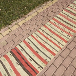 以下が含まれることがあります： 赤、茶色、クリーム色のストライプ模様の長方形のラグ。レンガの表面に緑の草の隣に置かれています。ラグのデザインは、縦縞と小さな装飾的な要素が特徴です。