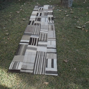 May include: A long, rectangular rug with a patchwork design. The rug is made of different shades of brown and white fabric, creating a geometric pattern. The rug is lying on a grassy surface.