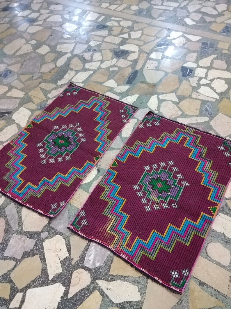 Puede incluir: Dos alfombras de color burdeos con patrones geom&eacute;tricos en tonos de azul, verde, rosa y amarillo. Las alfombras est&aacute;n sobre un suelo de baldosas.