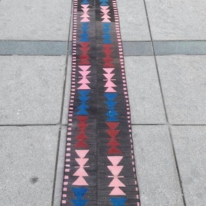 May include: A long, narrow rug with a geometric pattern in shades of pink, blue, red, and black. The rug has a fringed edge.