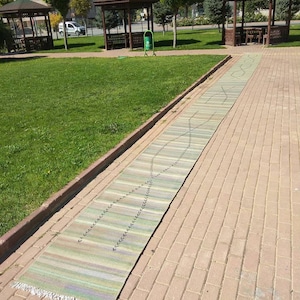 以下が含まれることがあります： 公園のレンガ敷きの小道に、フリンジの縁取りがついた長いストライプの敷物が敷かれている。敷物は小さな黒い点の模様があり、柔らかく織られた素材で作られている。