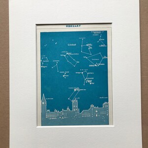 May include: A framed print showing a February constellation map in blue and white. The map displays star formations such as Gemini and Orion, with a cityscape silhouette at the base. The print is mounted in a white mount.