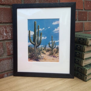 K&ouml;nnte beinhalten: Gerahmter Druck mit einer W&uuml;stenlandschaft mit Saguaro-Kakteen unter blauem Himmel mit Wolken. Der Druck ist in einem wei&szlig;en Passepartout und einem schwarzen Holzrahmen gefasst. Das Kunstwerk wird auf einer hellen Holzoberfl&auml;che pr&auml;sentiert.