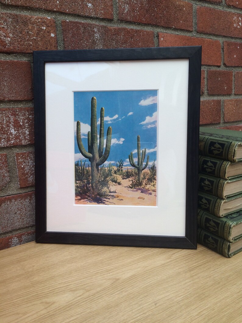 K&ouml;nnte beinhalten: Gerahmter Druck mit einer W&uuml;stenlandschaft mit Saguaro-Kakteen unter blauem Himmel mit Wolken. Der Druck ist in einem wei&szlig;en Passepartout und einem schwarzen Holzrahmen gefasst. Das Kunstwerk wird auf einer hellen Holzoberfl&auml;che pr&auml;sentiert.