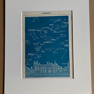 May include: A framed print showing a vintage January celestial map. The map is a deep blue with white constellations and text. The print includes a detailed illustration of a building at the bottom. The print is matted in white.