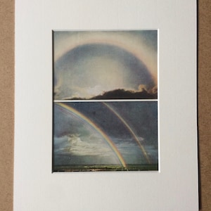May include: Framed artwork with two rainbow images against a cloudy sky. The top image shows a partial rainbow, the bottom a full rainbow over a landscape. The artwork is matted in white, creating a visual contrast.