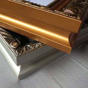 May include: Close-up of two ornate picture frames. The top frame is gold with a curved edge, while the bottom frame is silver with detailed corner carvings. Both frames have a classic, decorative style.