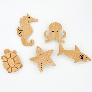 May include: Five wooden sea creature shapes, including a starfish, a seahorse, an octopus, a turtle, and a shark. The shapes are all made of light brown wood and have a natural finish.