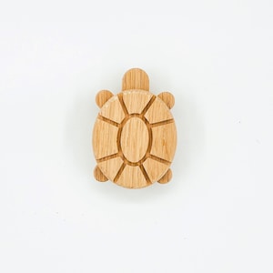 May include: A wooden turtle-shaped carving with a light brown finish. The turtle has a detailed shell design with radiating lines and small, rounded flippers. The head and flippers are also carved from wood.