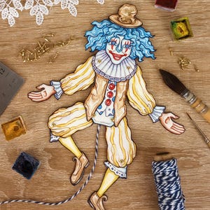 May include: A whimsical, hand-drawn clown puppet with blue hair, a small hat, and a colorful costume. The puppet is made of paper and has a string attached. Watercolor paints, a paintbrush, and other craft supplies surround the puppet on a wooden surface.