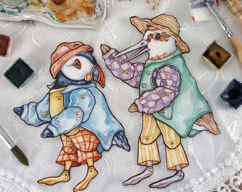 Puede incluir: Dos figuras de pájaros pintadas a mano con extremidades articuladas. Uno es un frailecillo con abrigo azul y pantalones a cuadros, el otro un pájaro con chaleco verde y mangas a cuadros. Ambos llevan sombreros y están adornados con cierres dorados.
