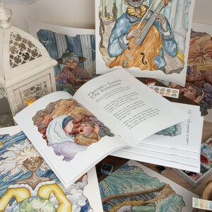 K&ouml;nnte beinhalten: Eine Sammlung von Aquarellbildern, die verschiedene Szenen darstellen, darunter ein Musiker, der Cello spielt, eine Frau, die einen Globus h&auml;lt, und eine Person, die in einem Boot schl&auml;ft. Die Bilder werden auf einem Tisch mit einem Buch mit dem Titel "Nativity Among The Suspects" ausgestellt, das auf einer Seite mit einer Aquarellillustration einer Frau und eines Kindes ge&ouml;ffnet ist.