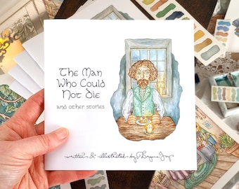 Könnte beinhalten: Ein quadratischer Buchumschlag mit dem Titel "The Man Who Could Not Die and other stories" und einer Illustration eines Mannes an einem Fenster. Der Umschlag wird von einer Hand gehalten, umgeben von anderen Kunstwerken und Aquarellzubehör.