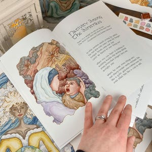 K&ouml;nnte beinhalten: Ein offenes Buch mit einer Aquarell-Illustration einer Frau, die ein weinendes Baby h&auml;lt. Das Buch tr&auml;gt den Titel "Nativity Among The Airstrikes" und enth&auml;lt ein Gedicht &uuml;ber die Weihnachtsgeschichte. Das Buch liegt auf einem Tisch mit anderen Aquarell-Illustrationen und einer Farbpalette.