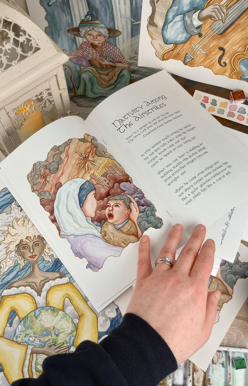 K&ouml;nnte beinhalten: Ein offenes Buch mit einer Aquarell-Illustration einer Frau, die ein weinendes Baby h&auml;lt. Das Buch tr&auml;gt den Titel "Nativity Among The Airstrikes" und enth&auml;lt ein Gedicht &uuml;ber die Weihnachtsgeschichte. Das Buch liegt auf einem Tisch mit anderen Aquarell-Illustrationen und einer Farbpalette.