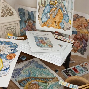K&ouml;nnte beinhalten: Ein Stapel Aquarellbilder und ein Buch mit dem Titel "Faces of God" auf einem Holztisch mit Pinseln und Aquarellpaletten. Die Bilder zeigen verschiedene Szenen, darunter einen Mann, der Cello spielt, eine Frau in einem Garten und einen Mann, der in einem Boot schl&auml;ft.