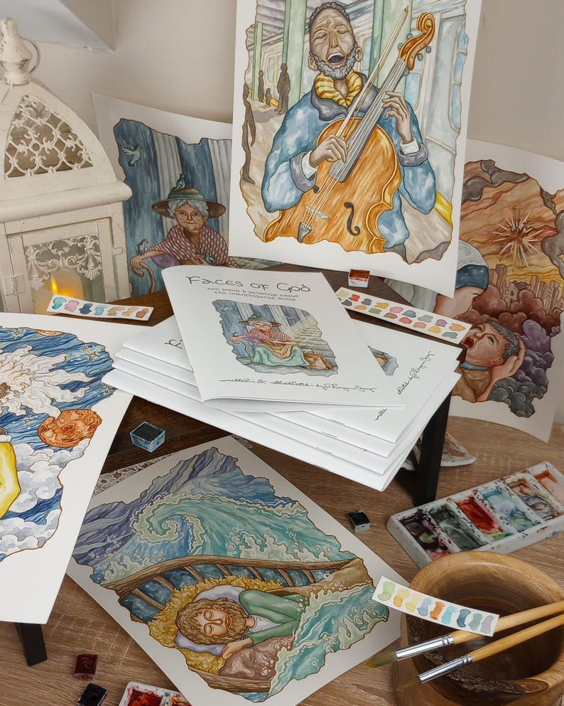 K&ouml;nnte beinhalten: Ein Stapel Aquarellbilder und ein Buch mit dem Titel "Faces of God" auf einem Holztisch mit Pinseln und Aquarellpaletten. Die Bilder zeigen verschiedene Szenen, darunter einen Mann, der Cello spielt, eine Frau in einem Garten und einen Mann, der in einem Boot schl&auml;ft.