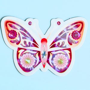 Op de afbeelding: Een gestanste sticker in de vorm van een vlinder met een holografische afwerking. Het ontwerp heeft symmetrische vleugels in roze, rode en witte tinten, versierd met bloemmotieven en een centraal stermotief op het lichaam, wat een reflecterend effect geeft.
