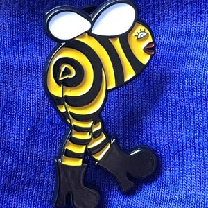 Può includere: Uno spillo in smalto nero e giallo di un'ape di cartone animato che indossa stivali neri e cammina. L'ape ha un viso bianco con una bocca rossa e ciglia.
