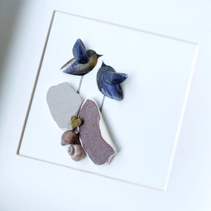 May include: A framed artwork featuring two blue birds made from seashells, perched on a brown and grey rock with a small gold heart.