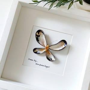 May include: A framed artwork featuring a butterfly made from seashells. The butterfly has white, black, and gold detailing. The artwork is in a white square frame, with the text "Dream big... then dream bigger!"