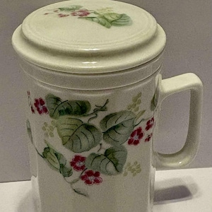 May include: Cream-coloured ceramic mug with a matching lid. The mug features a floral design with green leaves and red flowers. The mug has a handle and an octagonal shape.