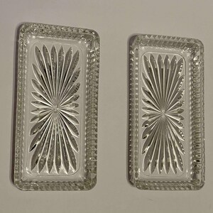 May include: Two clear glass rectangular dishes. Each dish features a starburst design and a textured border. These dishes are likely for serving or display, with a classic aesthetic.