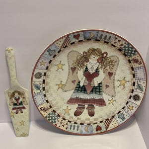 Peut inclure: Un ensemble décoratif comprenant une assiette de service et une pelle à tarte sur le thème des anges. L'assiette présente une illustration d'ange tenant un cœur, entourée d'étoiles et de cœurs. La pelle à tarte a un motif d'ange assorti.