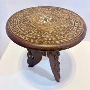 May include: A round, dark brown wooden side table with intricate floral carvings. The tabletop features a central inlaid design and is supported by three carved legs. The table is approximately 46 cm in diameter.