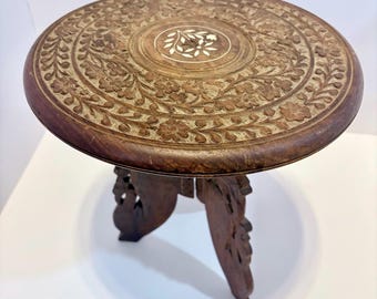 May include: A round, dark brown wooden side table with intricate floral carvings. The tabletop features a central inlaid design and is supported by three carved legs. The table is approximately 18 inches in diameter.